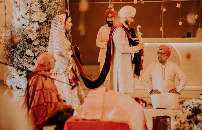 The Sikh bride stands next to her fiancé while the ceremony is ongoing. Both are wearing light beige clothes with red accessories.