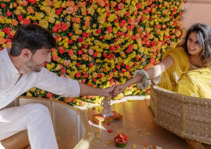 An Indian couple holding hands, surrounded by yellow, red, and orange flowers. They are both wearing traditional vests.