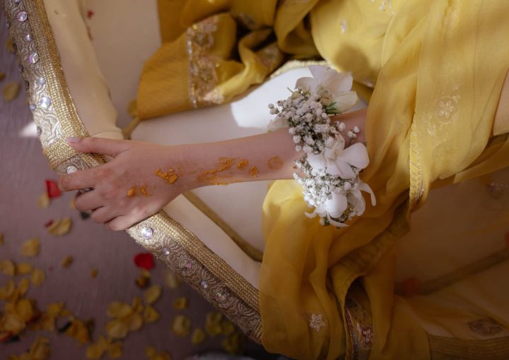 The arm of an Indian bride resting on the side of a chair. She has flowers on her elbow and is wearing yellow.