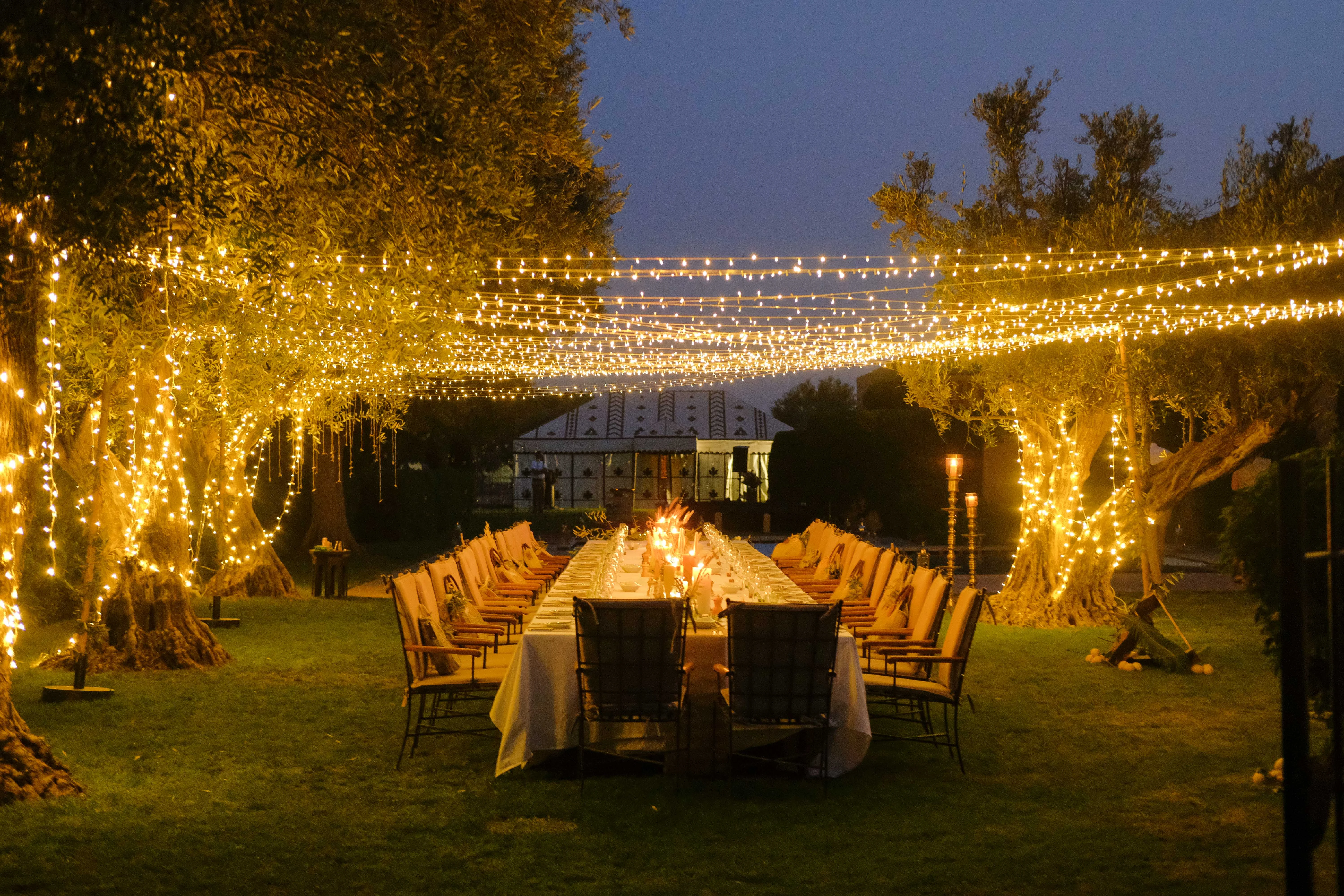 Long rectangular table in a garden with trees at night. The place is illuminated with candles and string lights.